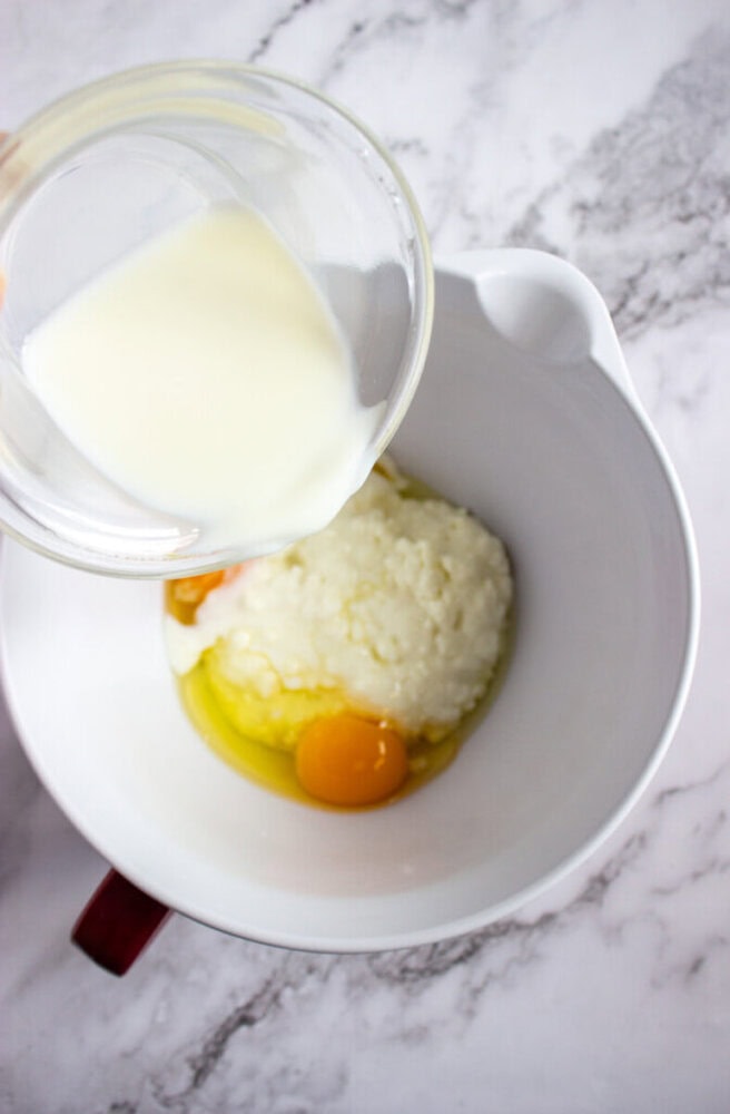 A glass measuring cup pours milk into a mixing bowl containing eggs and ricotta cheese on a marble countertop.