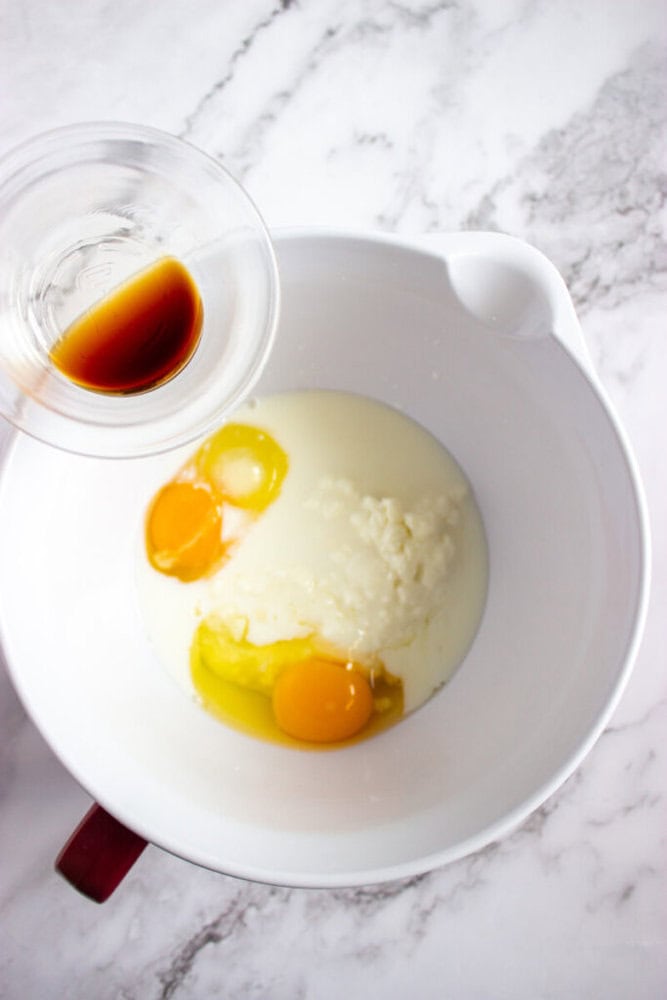 A bowl containing two eggs, milk, and ricotta cheese. A hand is pouring vanilla extract from a small cup into the bowl.