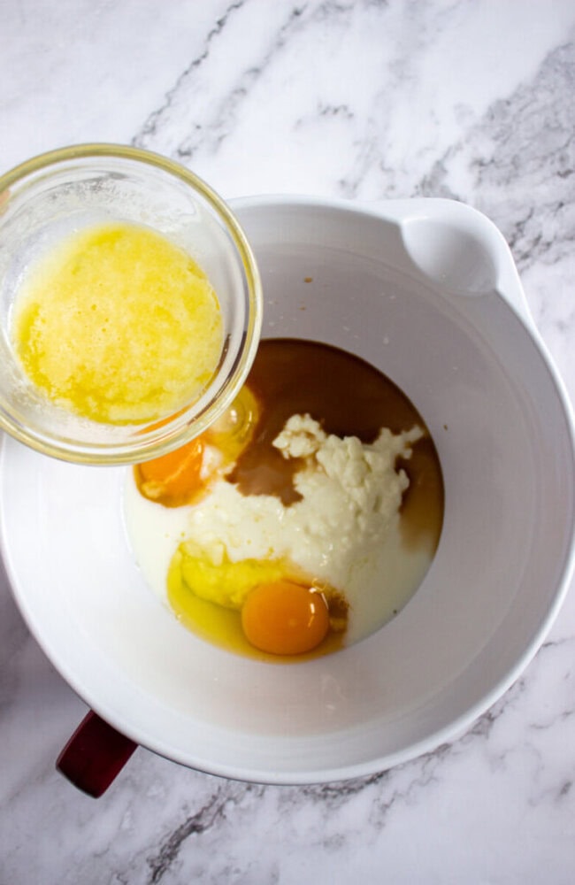 A bowl containing eggs, milk, and other ingredients, with melted butter being poured in from a glass container on a marble countertop.