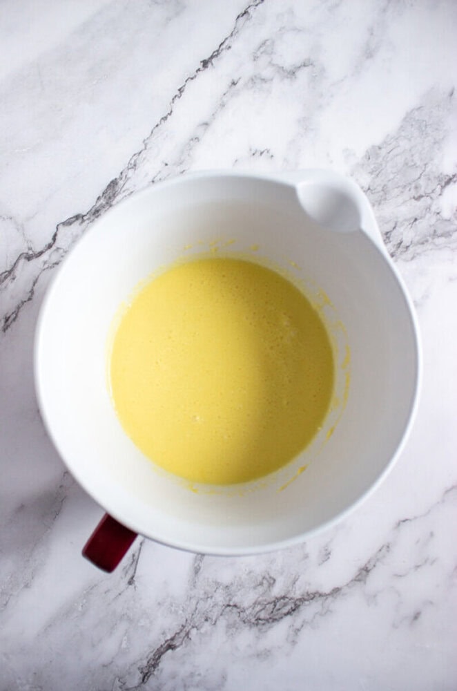 A white mixing bowl with a spout containing a smooth, light yellow batter sits on a white marble surface.