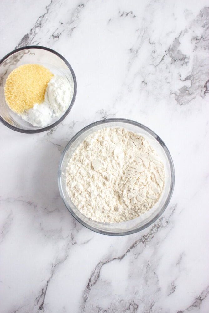 Two glass bowls on a marble surface, one containing flour and the other holding cornmeal and a white powder, possibly baking powder or baking soda.