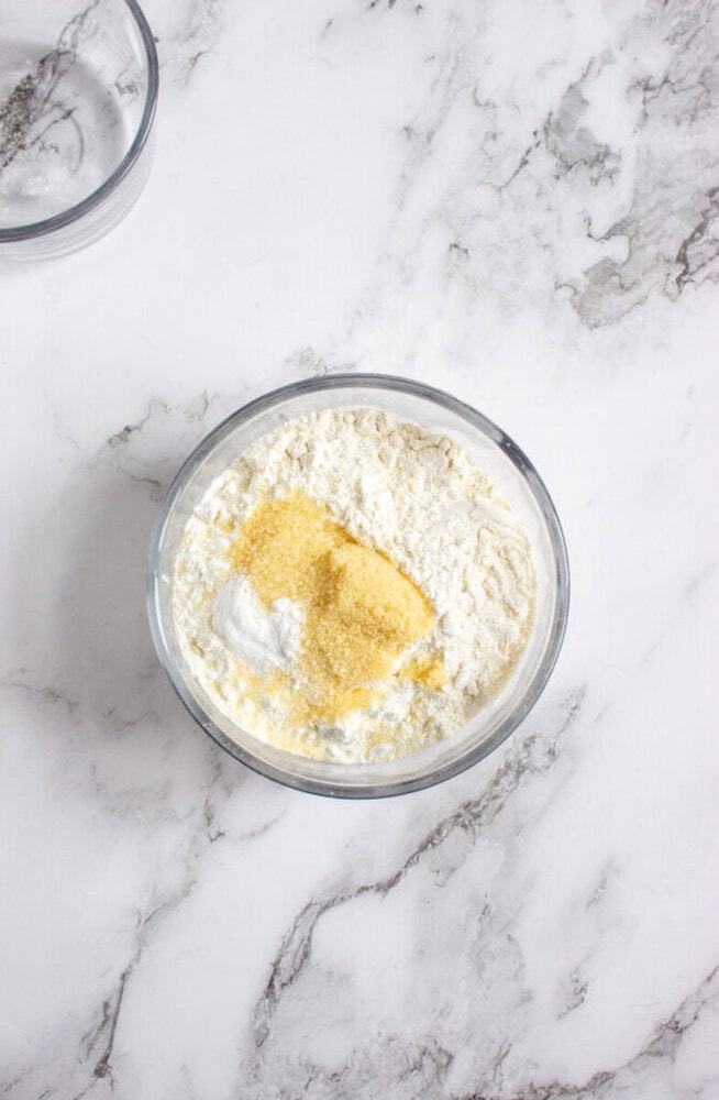 A clear glass bowl containing flour, cornmeal, baking soda, and other dry ingredients sits on a white marble surface.