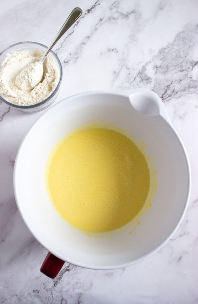 A large white mixing bowl with yellow batter and a smaller bowl of flour with a spoon on a marble countertop.