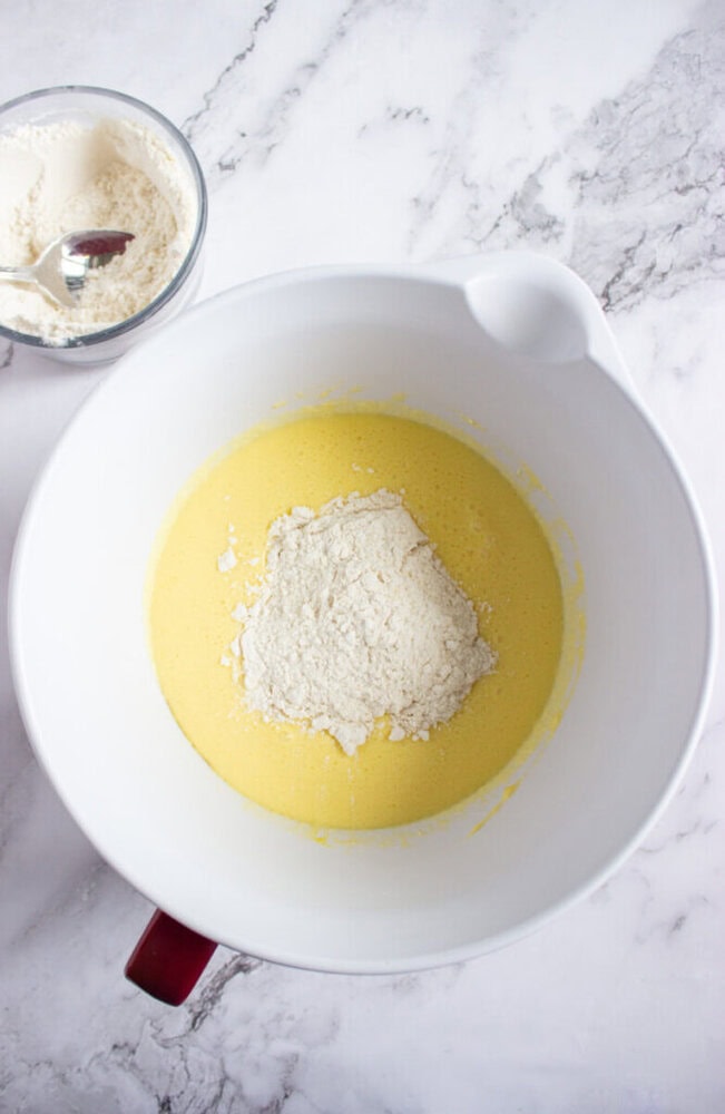A mixing bowl containing yellow batter with a mound of flour on top, next to a small bowl of flour on a white marble surface.