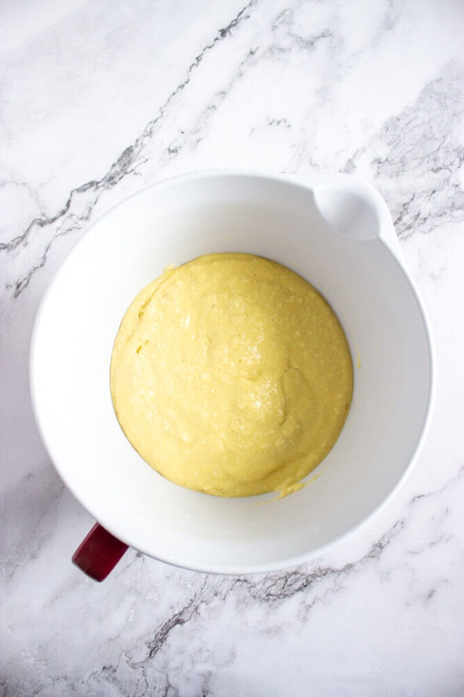 A white mixing bowl containing smooth, yellow batter sits on a white marble countertop.