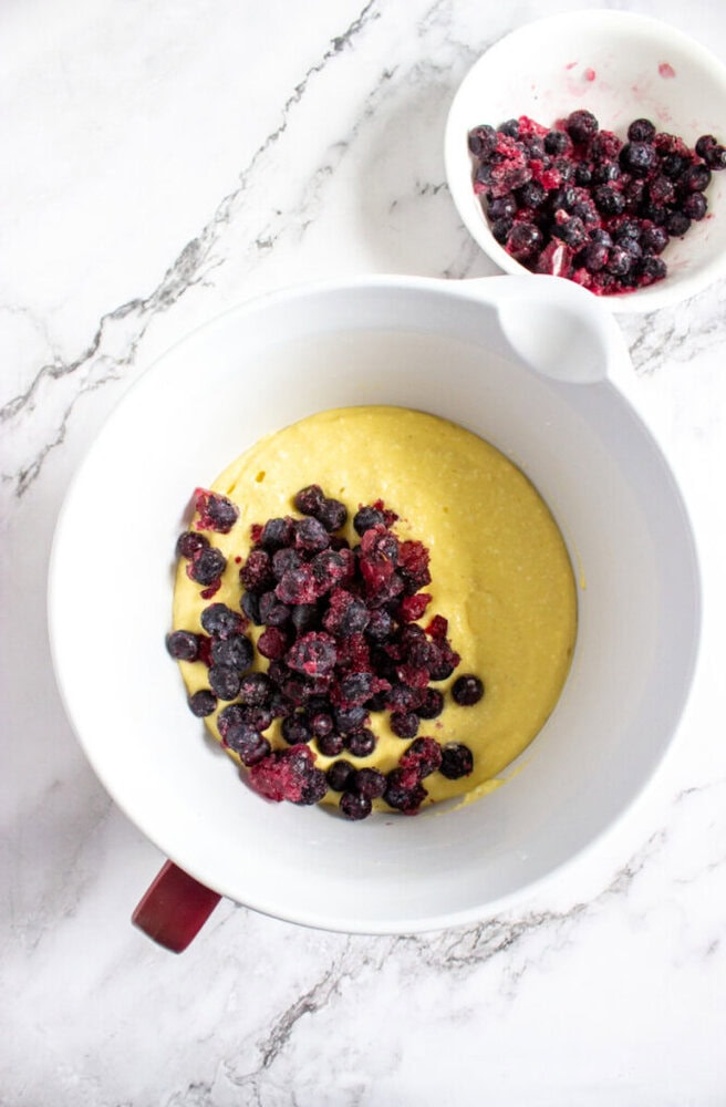 A white mixing bowl with yellow batter and frozen berries on top, next to a smaller bowl with additional frozen berries on a white marble surface.