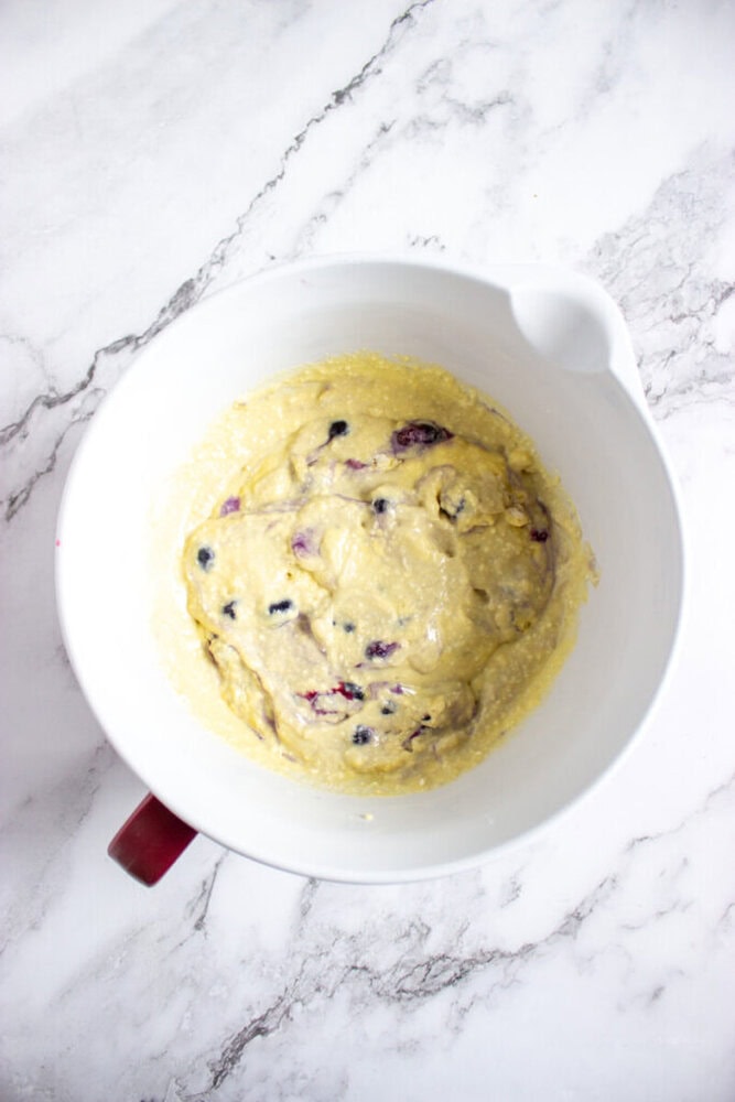A white mixing bowl containing thick, lumpy batter with visible blueberries, placed on a marble countertop.