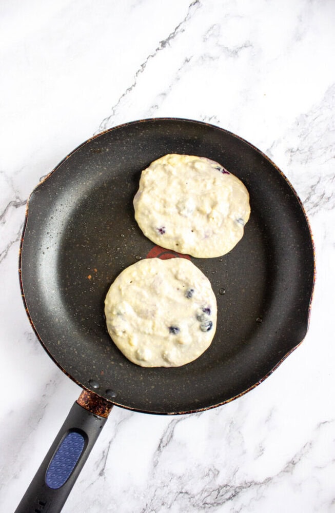 Two uncooked blueberry pancakes in a nonstick frying pan on a white marble surface.