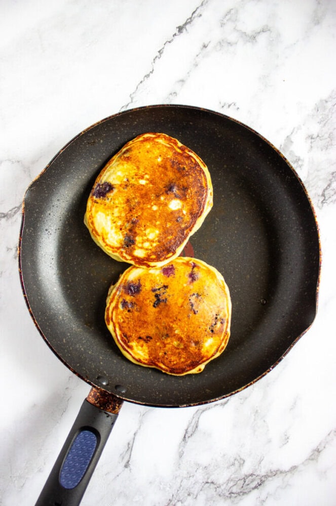 Two golden-brown pancakes with visible blueberries cook in a nonstick frying pan on a white marble surface.