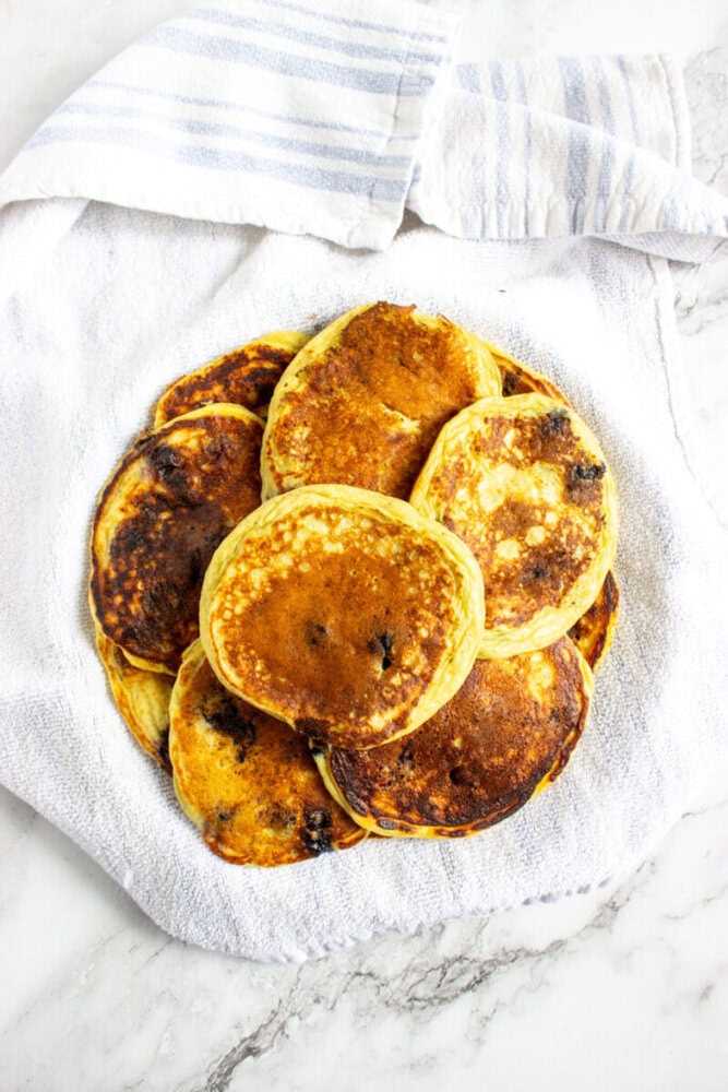 A plate of golden-brown pancakes is placed on a white towel with a striped cloth in the background, on a marble surface.