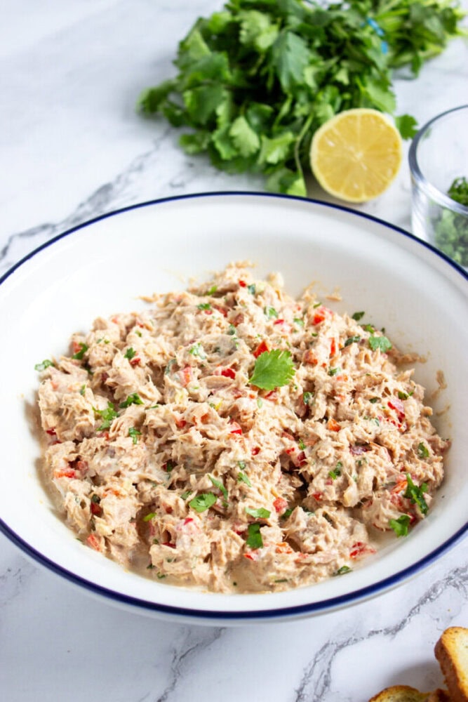 A white bowl filled with tuna salad mixed with chopped red peppers and herbs, placed on a marble surface with fresh cilantro and a halved lemon nearby.