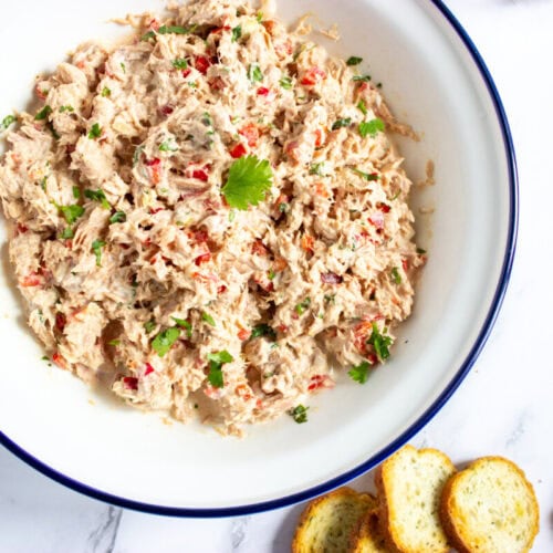 A bowl of tuna salad garnished with herbs sits on a white surface next to three slices of toasted baguette.