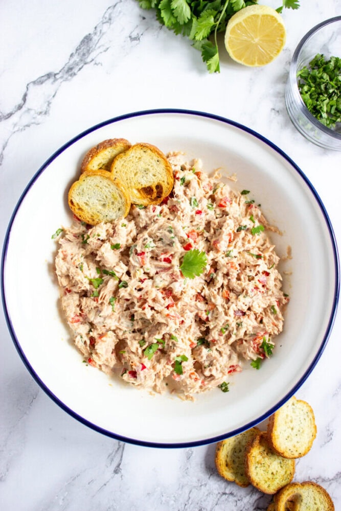 A white bowl filled with tuna salad garnished with cilantro and served with toasted bread slices, placed on a marble surface with herbs and a lemon nearby.