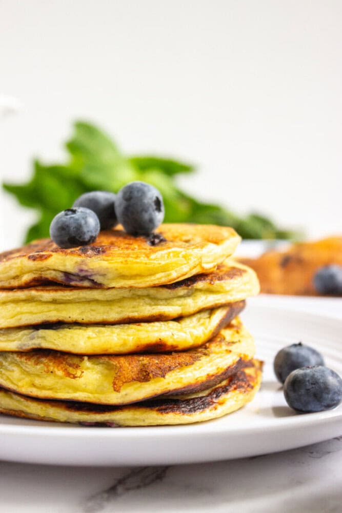 A stack of blueberry pancakes sits on a white plate, topped with fresh blueberries, with more blueberries and green leaves in the background.