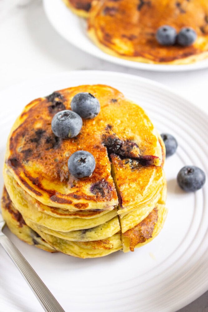 A stack of blueberry pancakes on a white plate, topped with fresh blueberries, with additional pancakes and berries in the background.