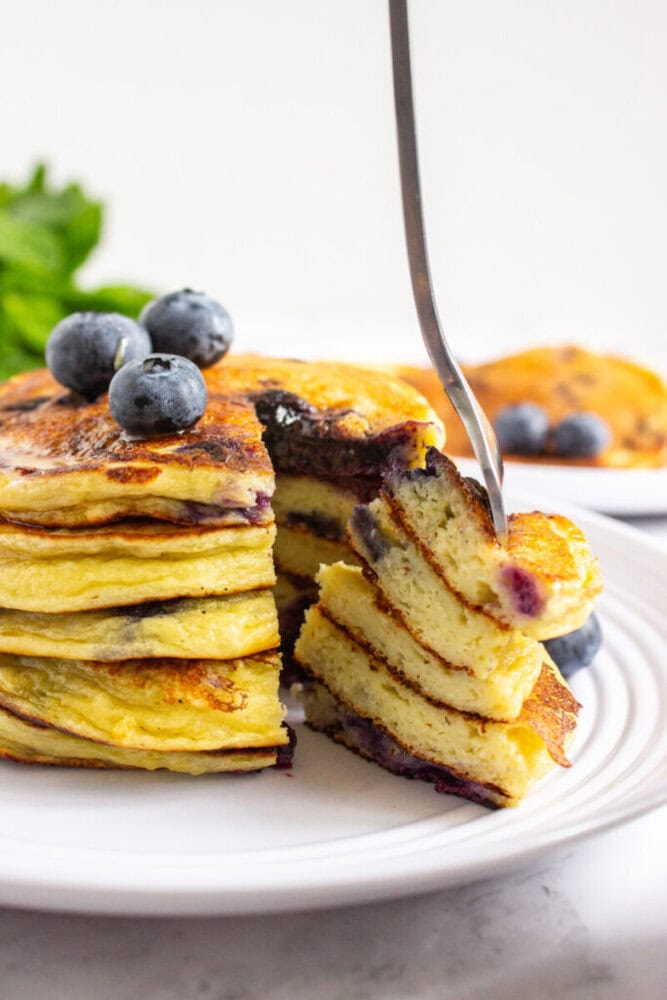 A stack of blueberry pancakes on a white plate, topped with fresh blueberries. A fork holds a bite-sized piece in front of the stack.