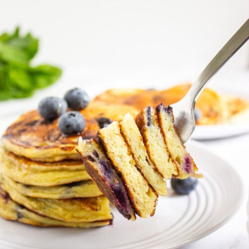 A fork holds a sliced stack of blueberry pancakes above a plate with more pancakes and blueberries, with some green leaves in the background.