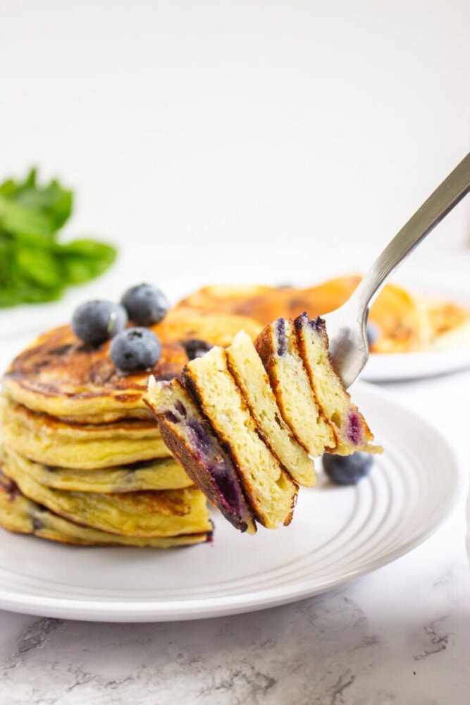 A fork holds a sliced stack of blueberry pancakes above a plate with more pancakes and blueberries, with some green leaves in the background.
