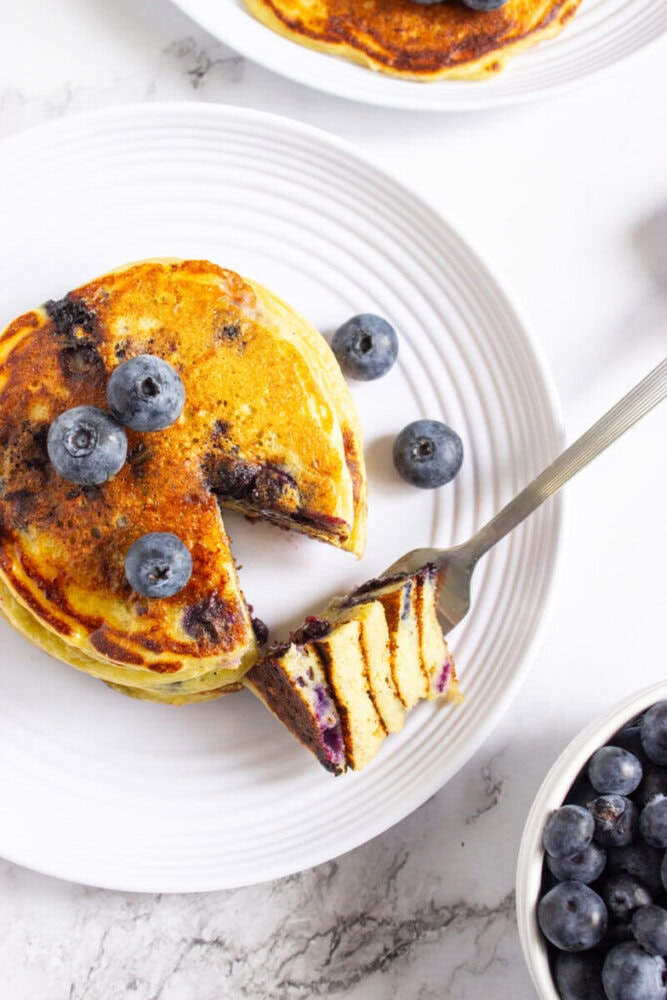 A stack of blueberry pancakes on a white plate, topped with fresh blueberries. A fork holds a cut slice, and extra blueberries are in a bowl nearby.