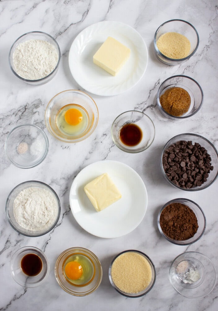 Top-down view of various baking ingredients in bowls and plates, including flour, eggs, butter, sugar, vanilla, chocolate chips, cocoa powder, and spices, arranged on a marble surface.