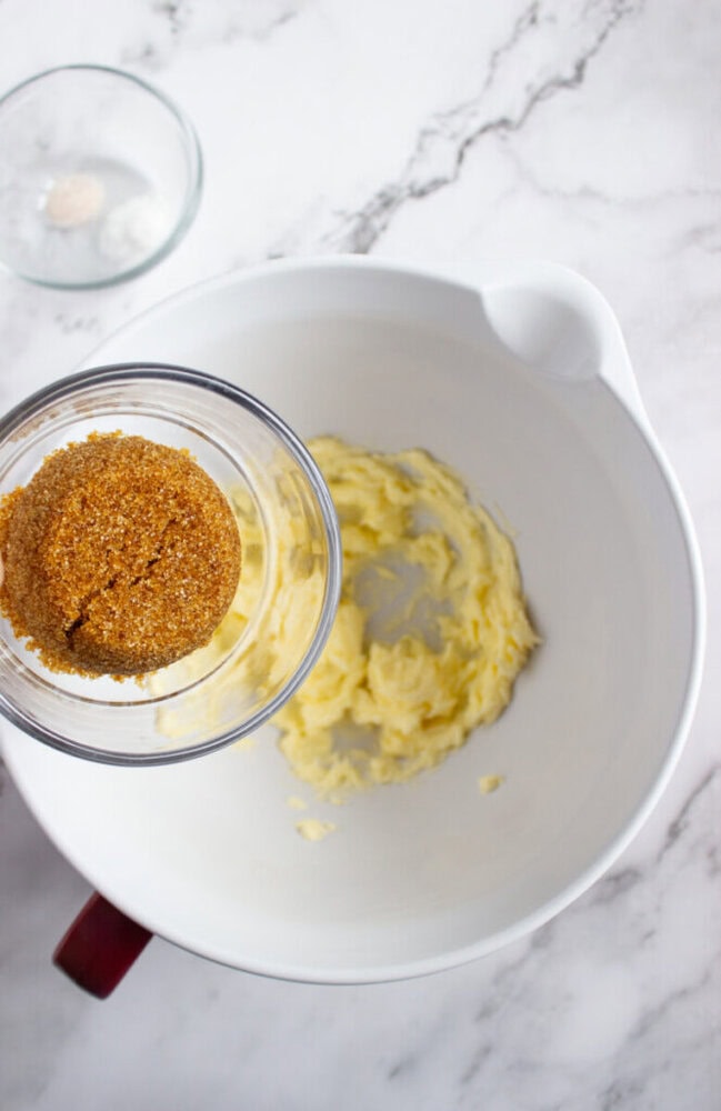 A glass bowl with brown sugar is held above a white mixing bowl containing creamed butter on a marble countertop.