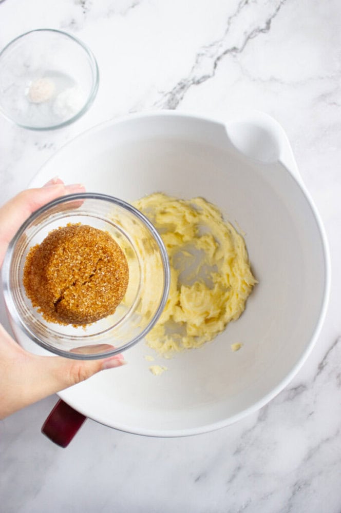 Hand holding a bowl of brown sugar over a mixing bowl containing creamed butter, on a marble countertop.