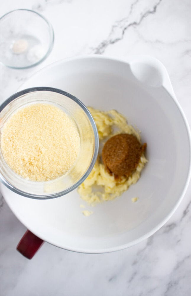 A bowl with mashed bananas and brown sugar sits on a marble counter as a glass container of granulated sugar is held above it, ready to be added.