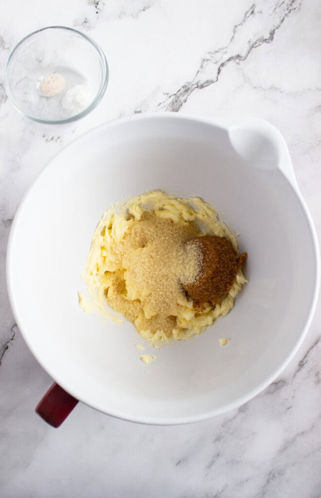 A white mixing bowl contains creamed butter and brown sugar on a marble countertop. A small glass bowl with salt and baking powder is nearby.