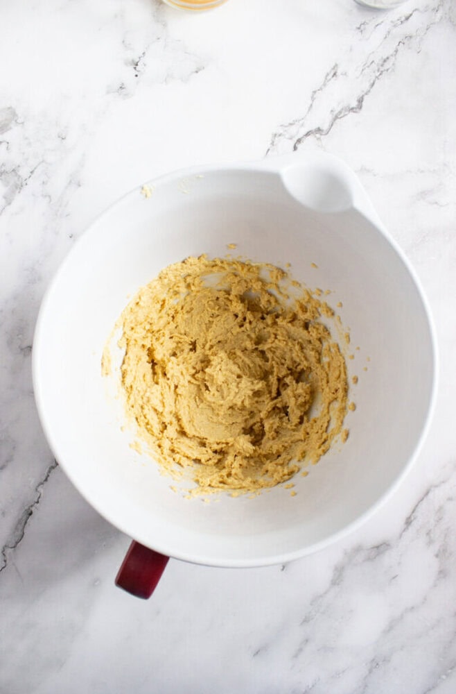 A white mixing bowl with tan cookie dough mixture sits on a marble countertop.