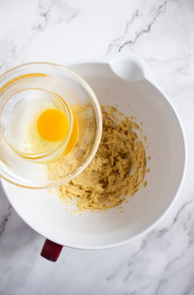 A glass bowl with a cracked egg is held above a mixing bowl containing partially mixed dough on a marble countertop.