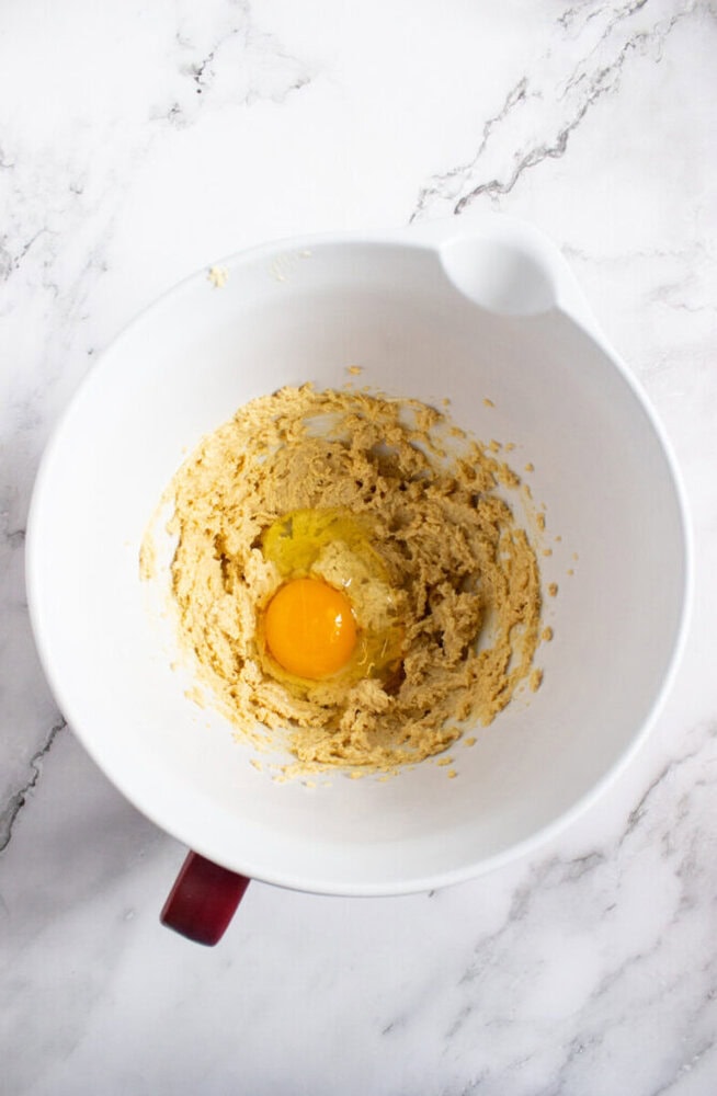 A white mixing bowl with cookie dough and a raw egg yolk in the center, placed on a marble countertop.