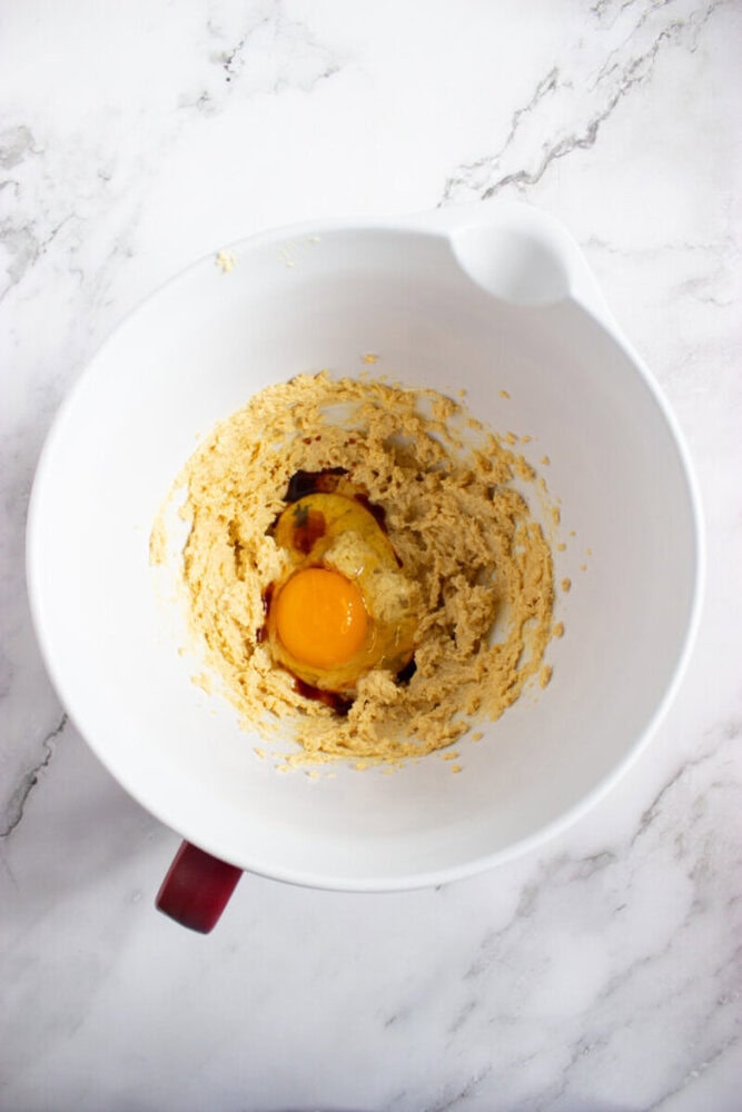 A mixing bowl containing creamed butter and sugar with a raw egg and vanilla extract on top, ready to be mixed, placed on a marble countertop.