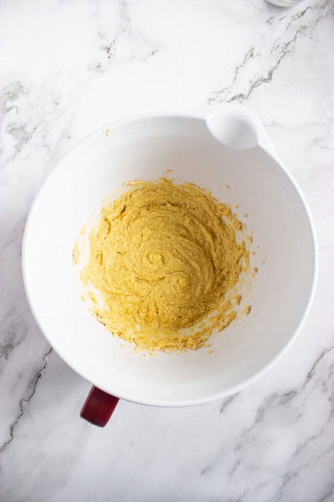 A white mixing bowl with a handle contains a thick, yellow batter on a white marble countertop.
