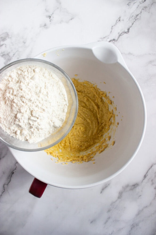 A bowl of flour is being added to a mixing bowl containing a yellow batter, on a white marble countertop.