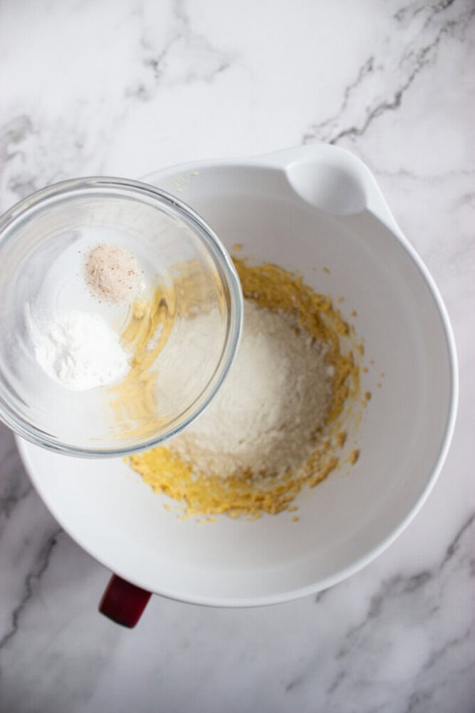 A glass bowl with salt and baking powder is being added to a mixing bowl containing flour and a yellow batter on a marble surface.