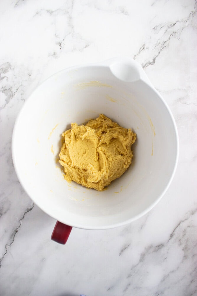 A white mixing bowl with a handle contains yellow cookie dough, placed on a white marble surface.