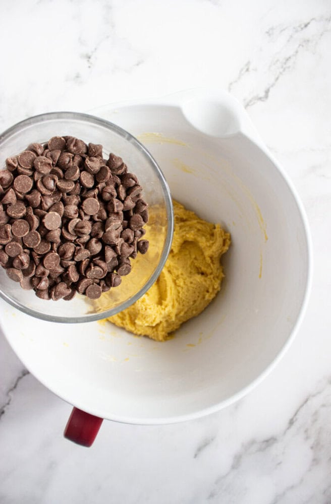 A glass bowl of chocolate chips is about to be poured into a mixing bowl containing cookie dough on a marble countertop.