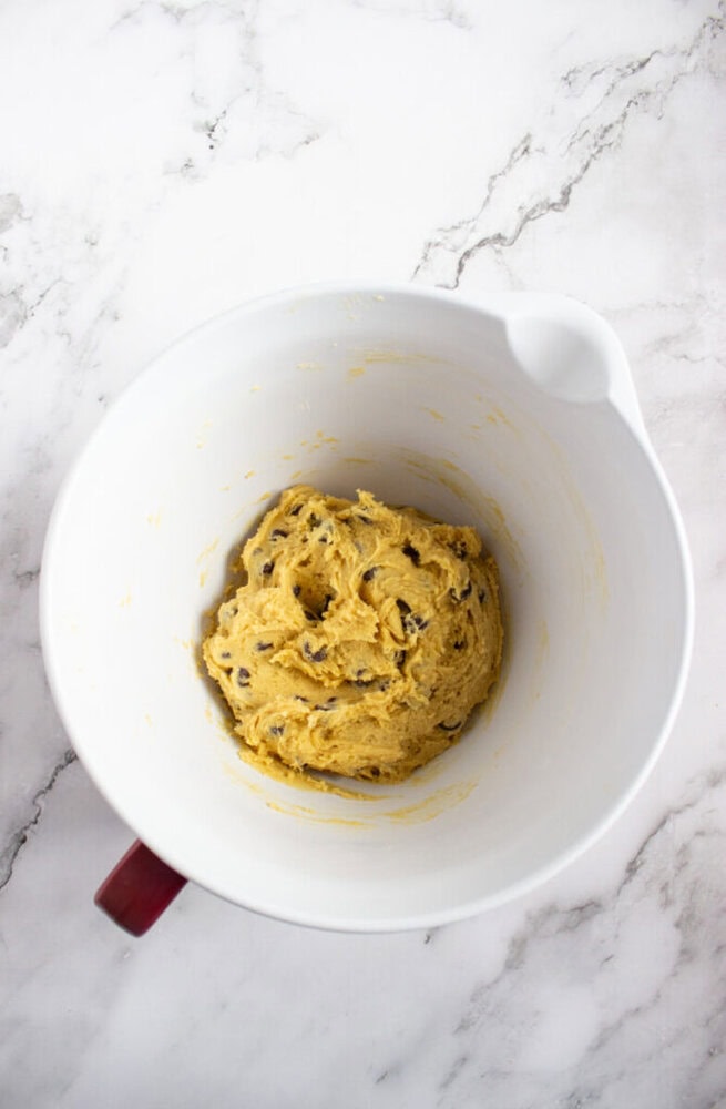 A white mixing bowl containing chocolate chip cookie dough sits on a marble countertop.