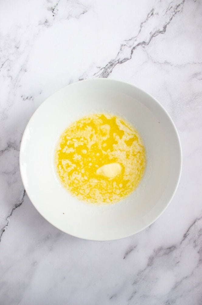 A white bowl containing melted butter sits on a white marble surface.