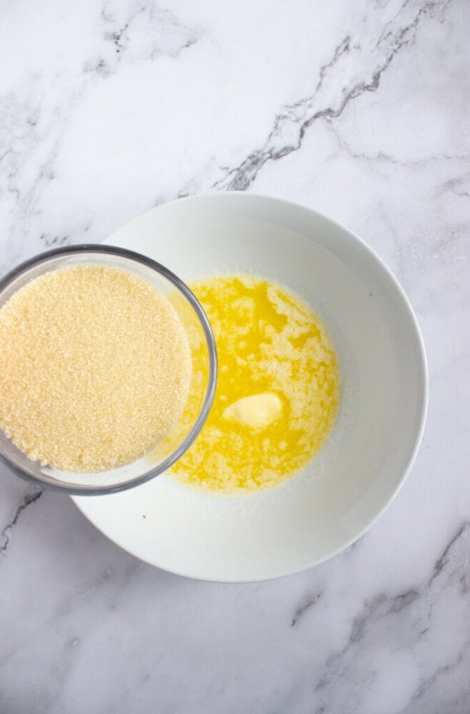 A bowl of melted butter sits on a marble surface next to a glass bowl filled with granulated sugar.