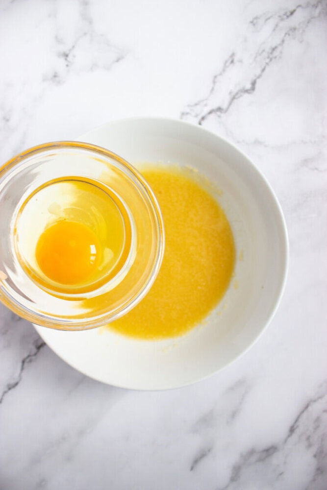 A glass bowl with a raw egg is held above a white bowl containing melted butter, all on a white marble surface.
