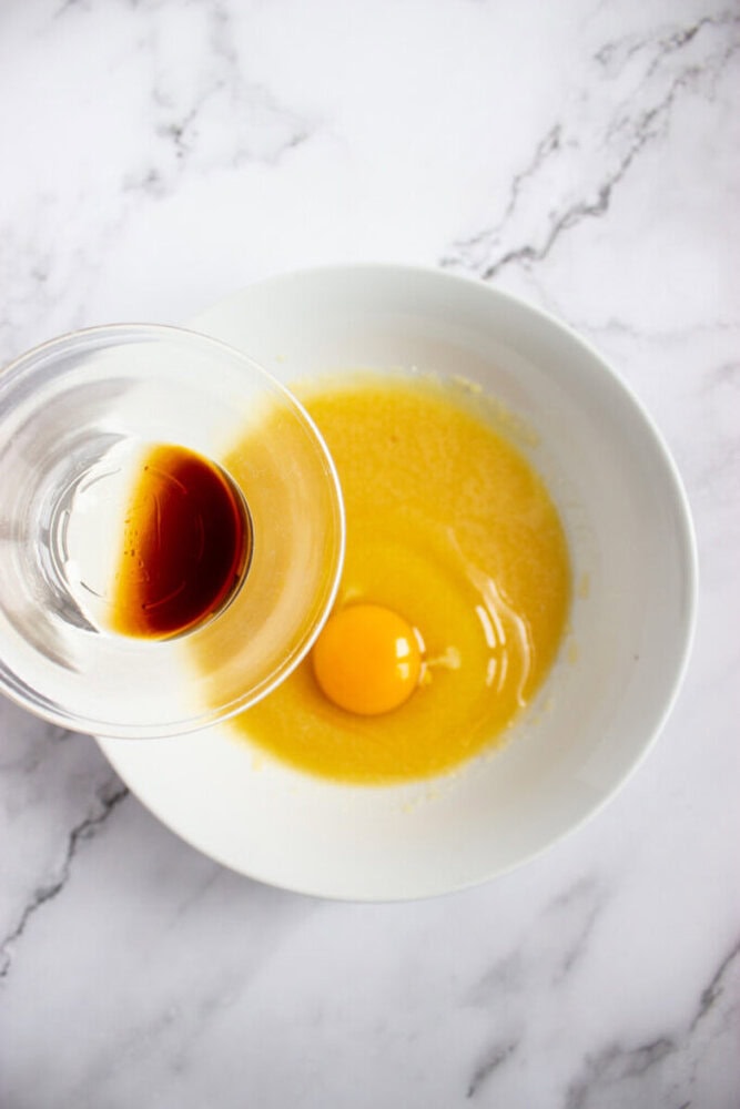 A glass bowl of vanilla extract held above a white bowl containing a raw egg and melted butter on a marble countertop.