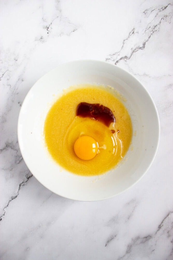 A white bowl contains a raw egg, brown vanilla extract, and beaten yellow butter-sugar mixture on a white marble surface.