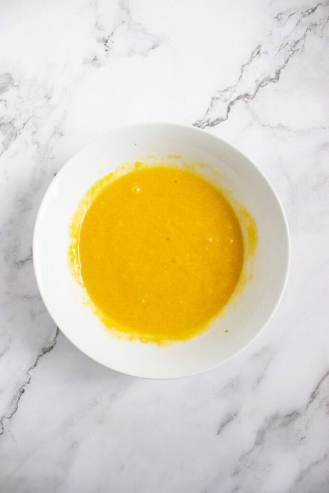 A white bowl containing a yellow liquid mixture sits on a white marble surface.