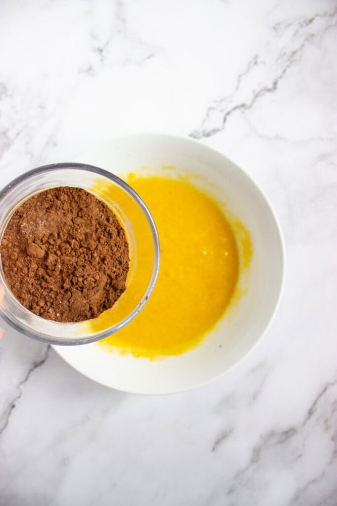 A bowl of yellow liquid sits on a marble surface while cocoa powder is being poured in from a measuring cup above.