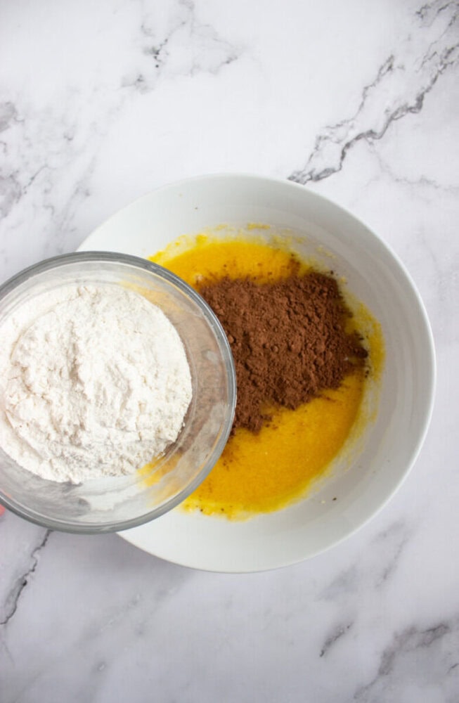 A bowl with eggs and melted butter is being mixed with flour and cocoa powder on a marble surface.