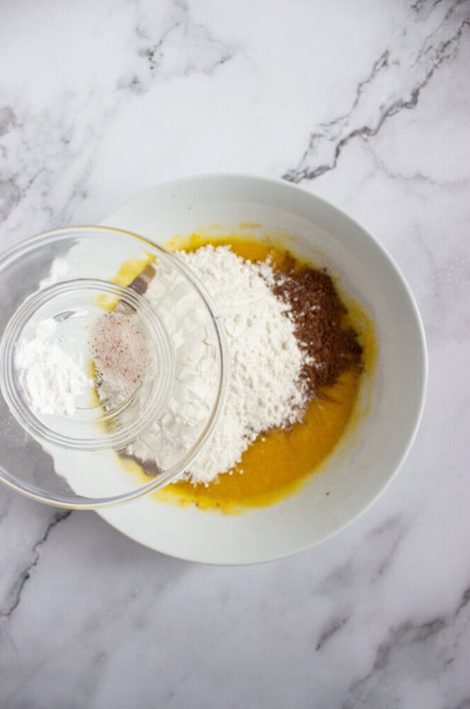 A mixing bowl with flour, cocoa powder, and wet ingredients, with a small bowl of salt and baking powder being added on a marble countertop.