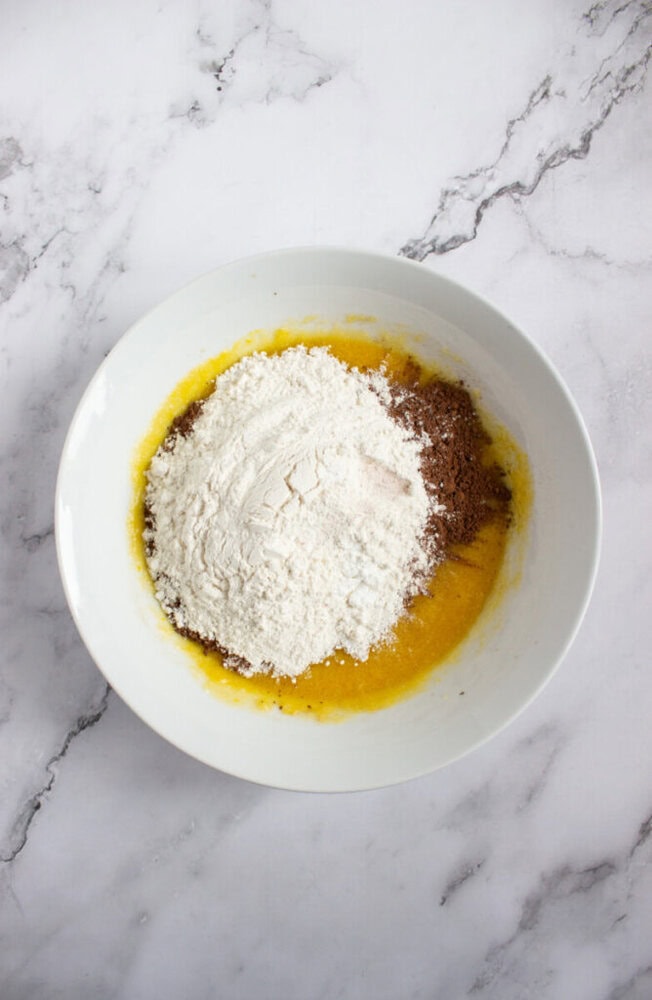 A white bowl on a marble surface contains flour, cocoa powder, and a yellow liquid mixture, ready to be mixed.