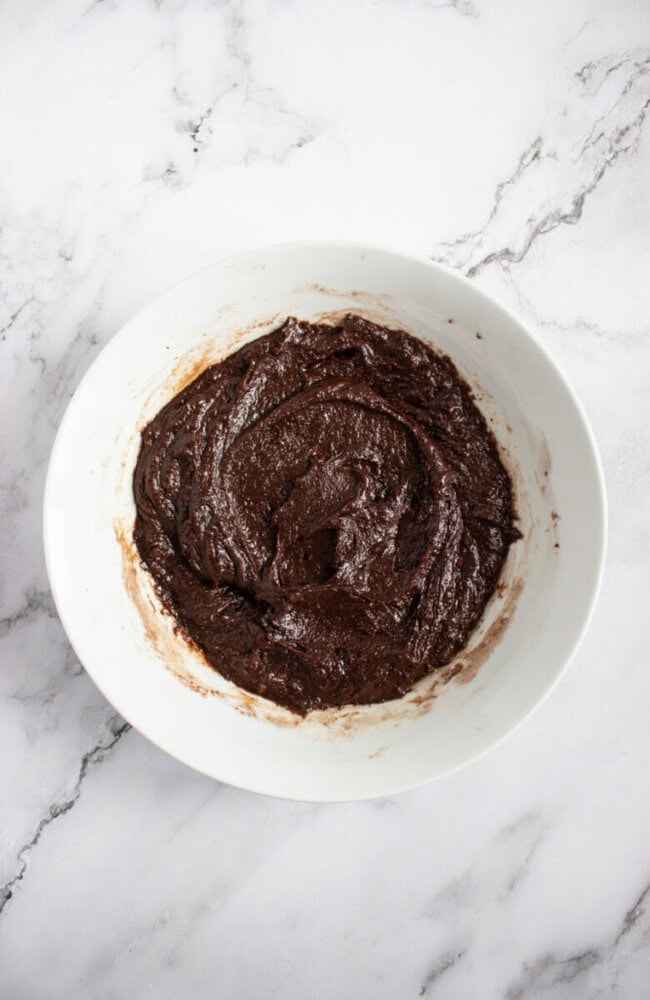 A white bowl filled with chocolate brownie batter sits on a white marble surface.