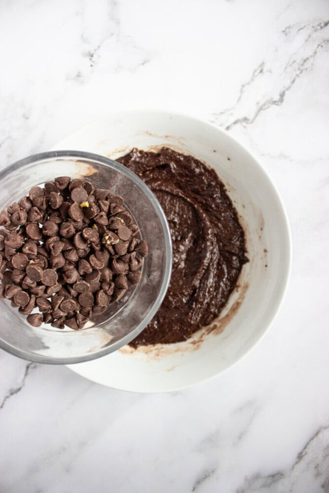 A bowl of chocolate chips is being held above a bowl of chocolate batter on a white marble surface.